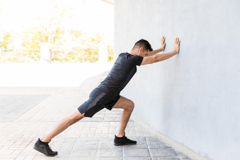 Determined young Latin man stretching calf while leaning on wall