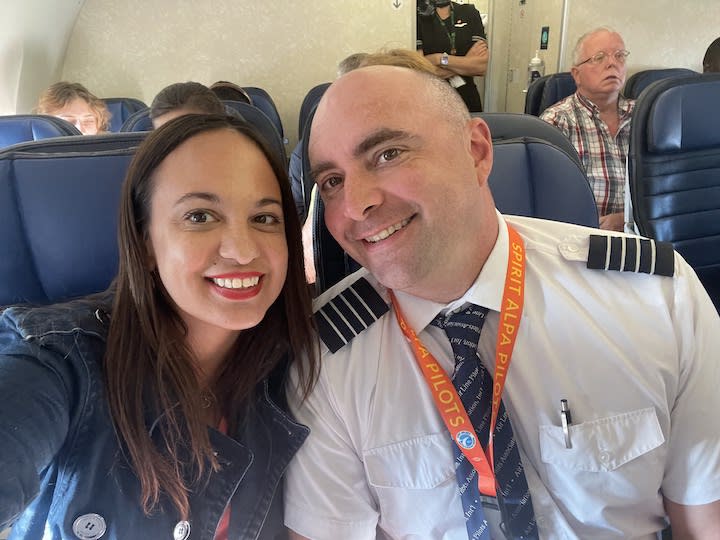 Two people sit in an airplane cabin. The man on the right wears a pilot outfit. 