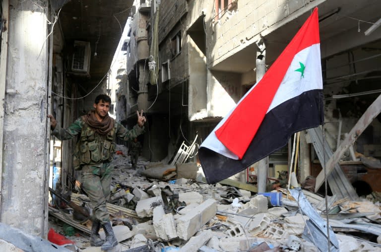 A member of the Syrian government forces poses for a photo next to a Syrian flag in destroyed buildings at the entrance of the Palestinian camp of Yarmuk on the southern outskirts of Damascus on May 21, 2018