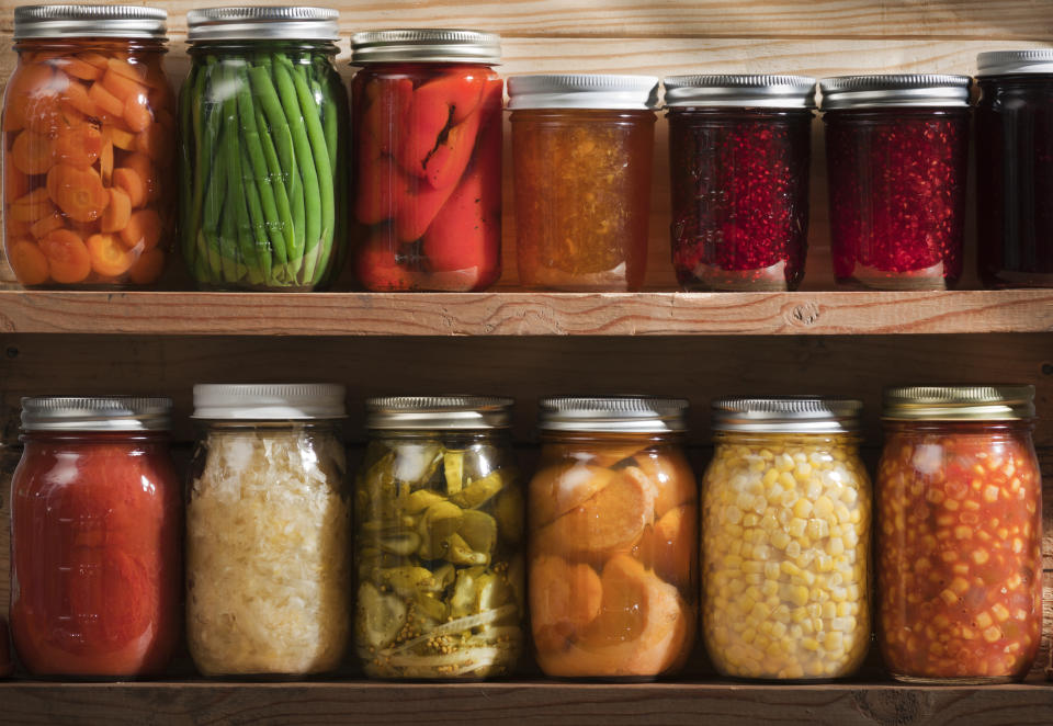 Two wooden shelves holding a variety of canned vegetables and fruits, lined up in rows of glass jars. Food staples canned include jellies, sauces, or slices of carrots, green beans, tomatoes, corn, sweet potatoes, sauerkraut, roasted red peppers, dill pickles, raspberry jam, orange marmalade, grape jelly, and a tomato and corn soup.