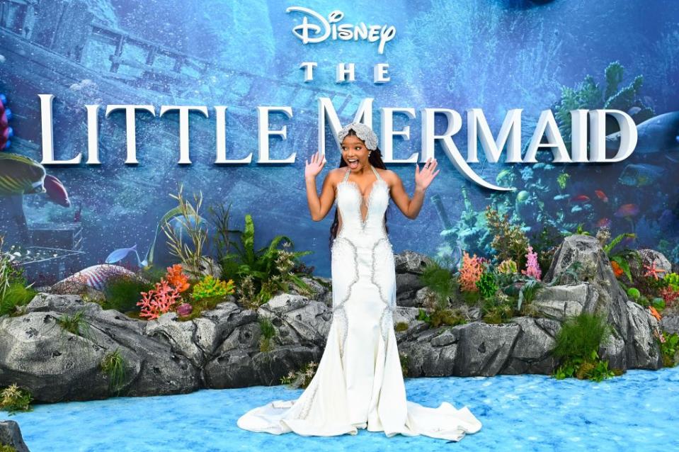 halle bailey stands in a white sleeveless gown in front of a photo background themed for the little mermaid