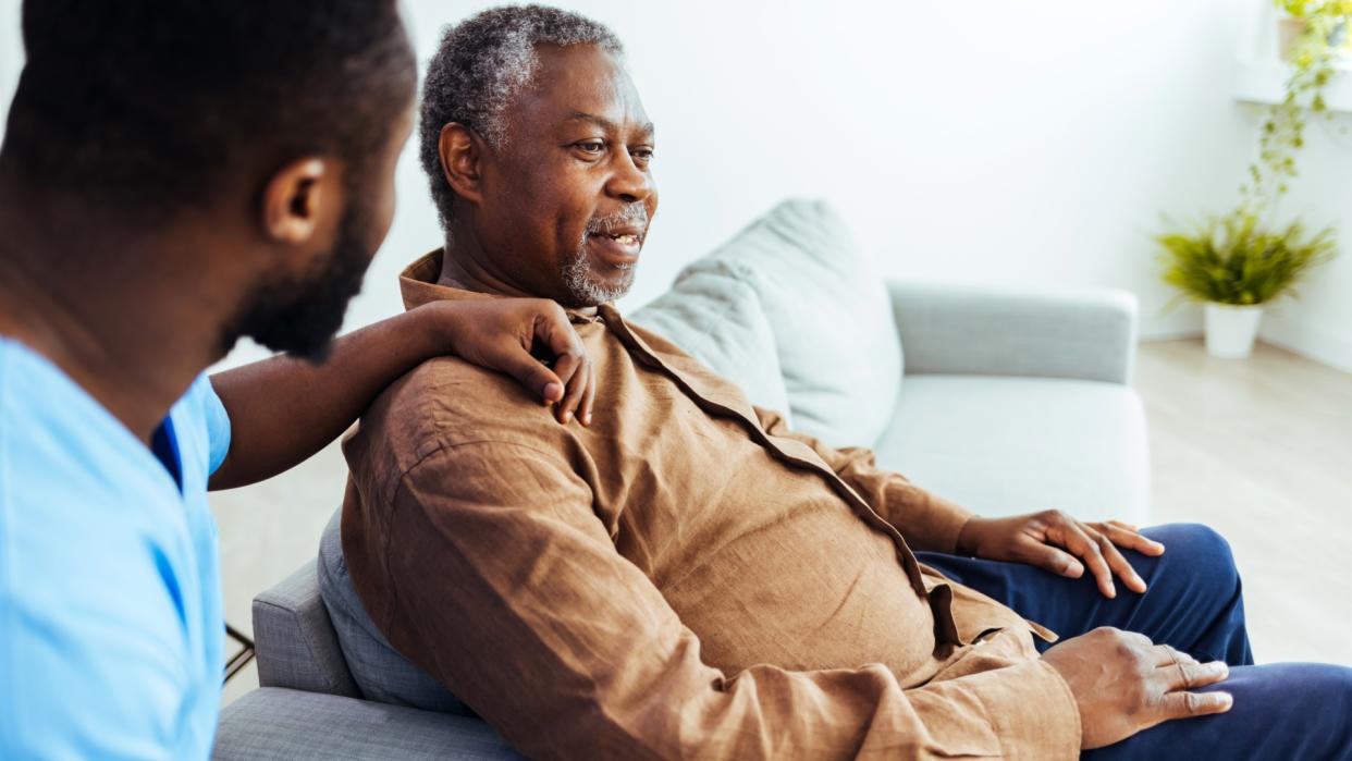  Social worker in foreground rests arm on shoulder of senior man who is sitting on a couch. 