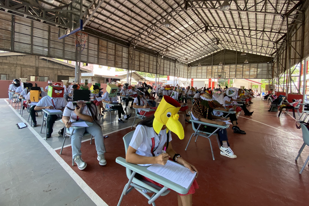 Students in a hall taking an exam, one wearing a chicken head mask, others in varied hats