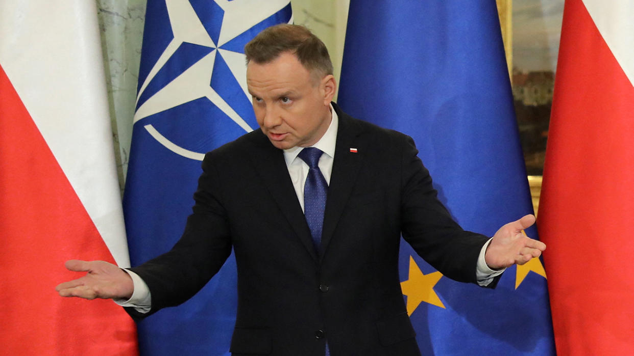 Polish President Andrzej Duda stands with arms extended and palms face up in front of a few flags.