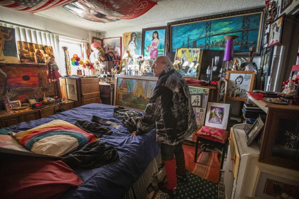 A man stands alongside a bed with a rainbow pillow in a room packed with photos and framed artwork.