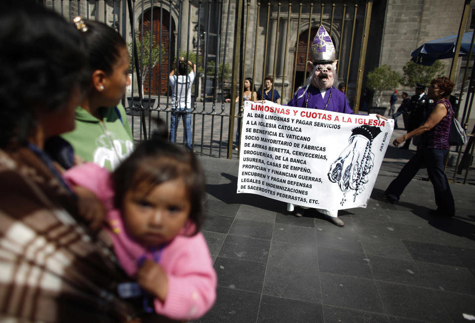A woman, dressed as a cardinal and wearing a hog mask, holds a banner while yelling slogans against the Catholic clergy outside the Metropolitan Cathedral in Mexico City
