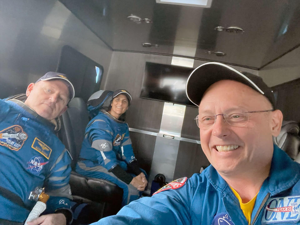 three astronauts in blue flight suits smile inside a gray van