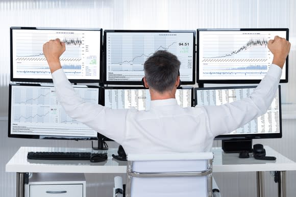 A man, seen from the back and seated at a desk, raises his clenched hands in a victory gesture as he looks at a bank of computer monitors.