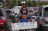 FILE - In this April 26, 2021 file photo, Jeferson Alves, 35, carries his 3-year-old son Jonathan Miguel on his shoulders when asking for a handout, as he holds a sign written in Portuguese that reads "Help me buy a basic food basket. Thank you," on an avenue in Brasilia, Brazil. Alves says he worked at a construction company for two years and that at the end of last year he was fired because of the pandemic. Now he seeks donations and money on the streets to support his family. (AP Photo/Eraldo Peres, File)