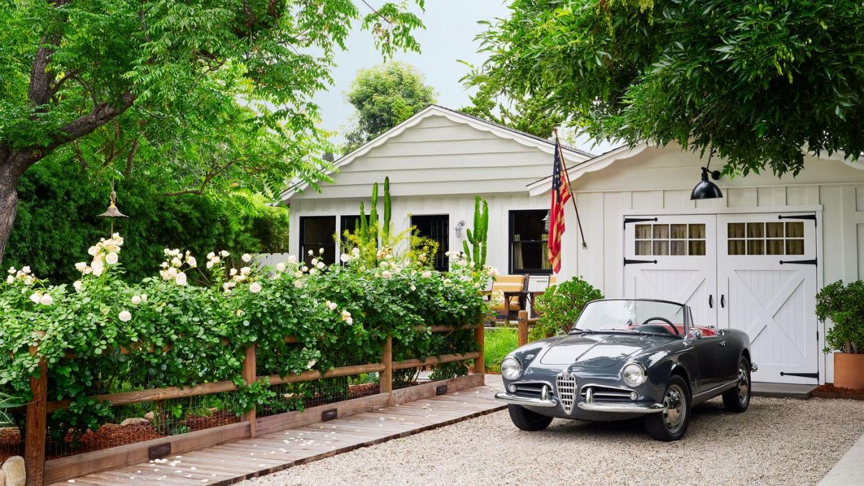 an alfa romeo spider convertible in a gravel driveway in front of a white garage with manual doors, a flag, walkway next to a fence with flowering rose bushes, white ranch house with windows, mature trees