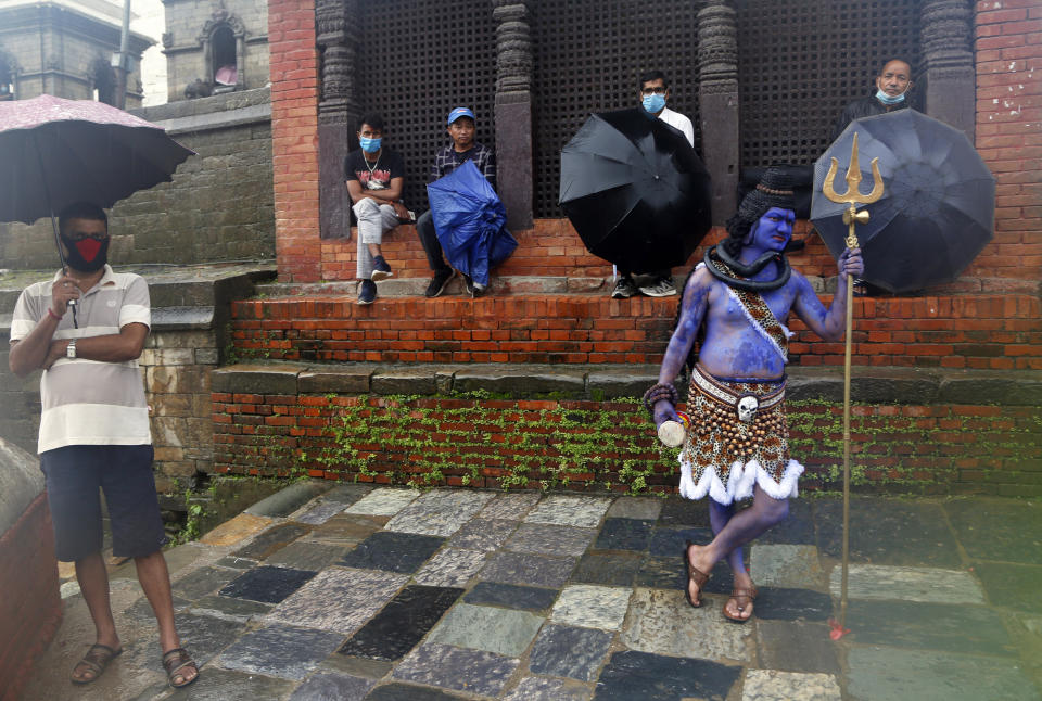 A member of a television crew stands dressed as Hindu god Shiva as Nepalese Hindu devotees wearing masks sit after offering prayers from outside the closed gate of Pashupatinath temple during the holy month of Shrawan in Kathmandu, Nepal, Monday, July 20, 2020. The temple has been remaining closed for almost four months as part of measures to control the spread of the coronavirus. (AP Photo/NIranjan Shrestha)
