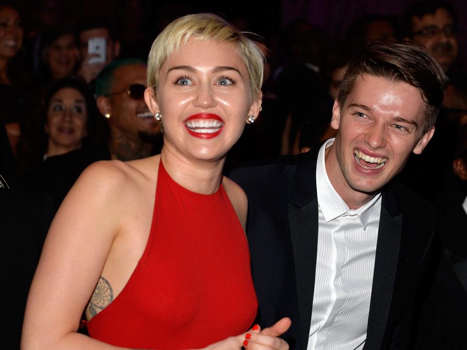 Miley Cyrus, in a red gown and lipsticks, smiles while at an event with Patrick Schwarzenegger, wearing a black suit and white shirt.