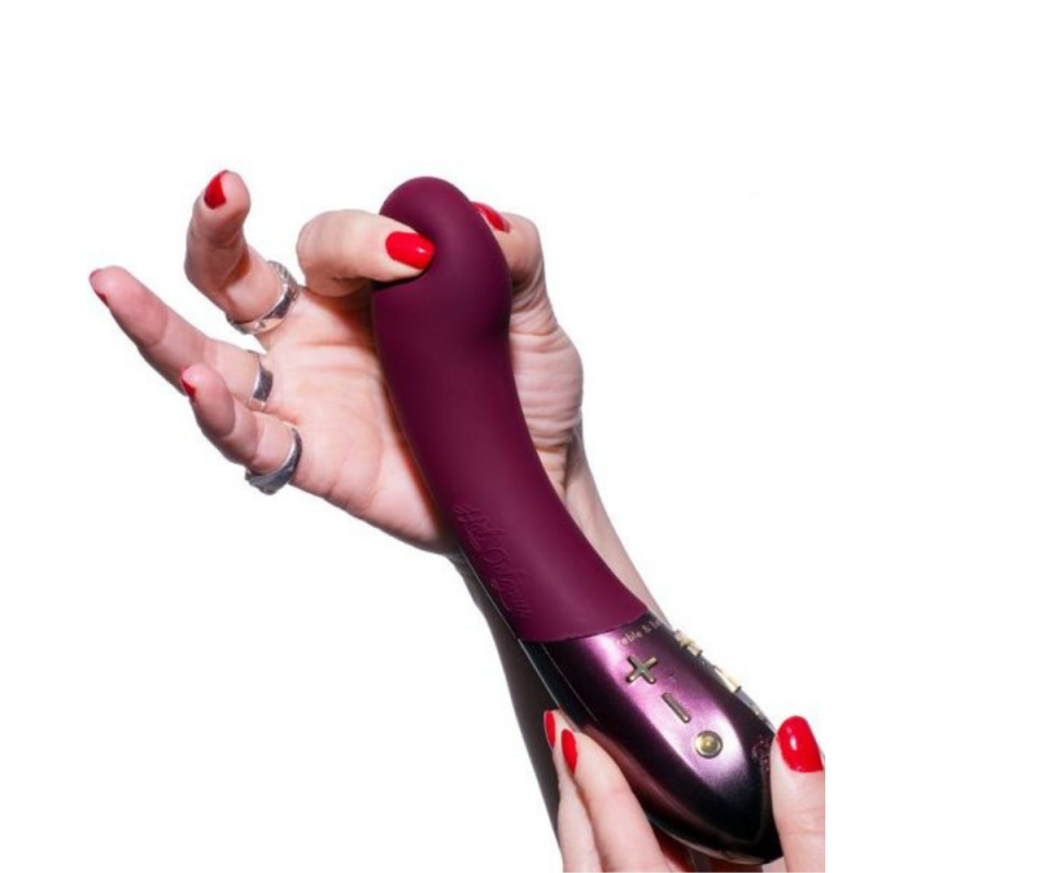 A caucasian woman's hand with red painted nails squishes the soft gel tip of a maroon vibrator against a white background