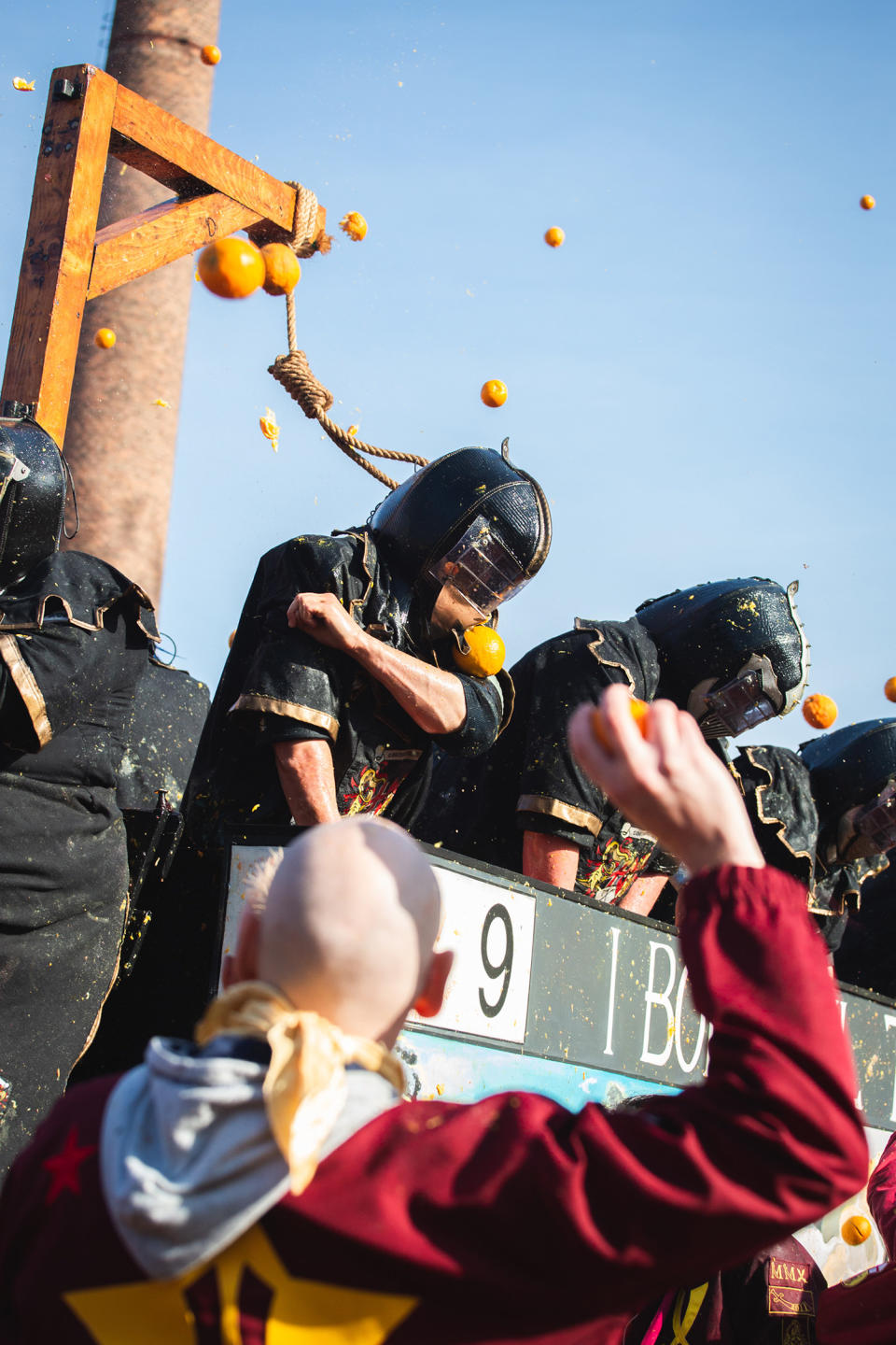 Standing under a noose, people wearing helmets on a raised platform are hit with oranges; in the foreground, a blurry man has his arm raised and an orange in his fist, about to chuck it at someone on the platform directly before him