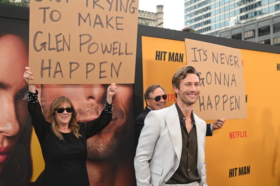 Glen Powell, a man in a white suit, smiles at an event. Two people hold signs saying "Stop trying to make Glen Powell happen" and "It's never gonna happen."