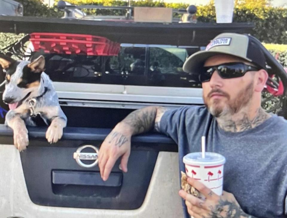 A man wearing sunglasses and a baseball cap holds a drink in his hand and leans an armon the bed of a pickup truck. A white dog with black patches on its eyes hangs over the edge of the truck bed.