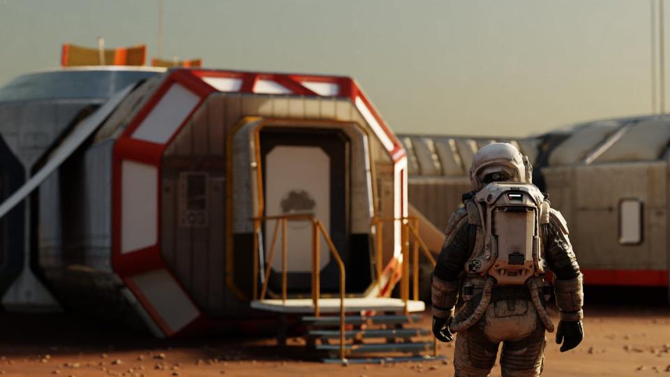 The image shows an astronaut on Mars standing in front of a red, white, and silver modular habitat.