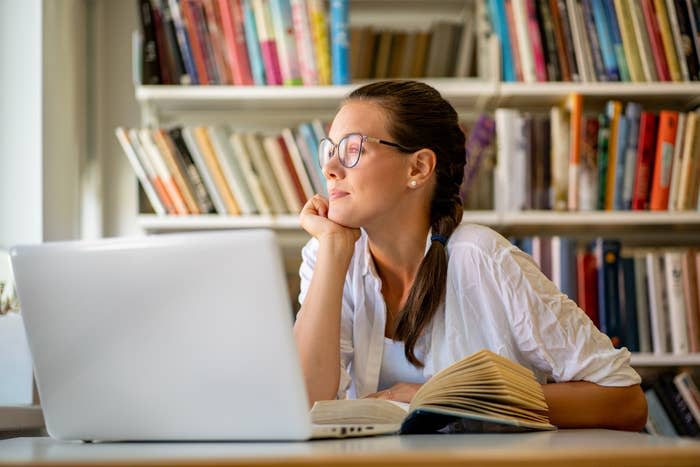 A woman sits at a desk, looking thoughtfully out a window, with an open book in front of her and a laptop. She is in a room with bookshelves filled with books