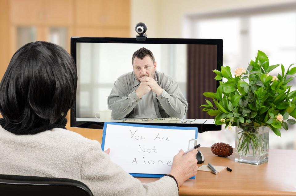 Man talks with psychotherapist via online video chat. He looking depressed. Black-haired psychiatrist holds written message for him - You are not alone. Horizontal shot indoors