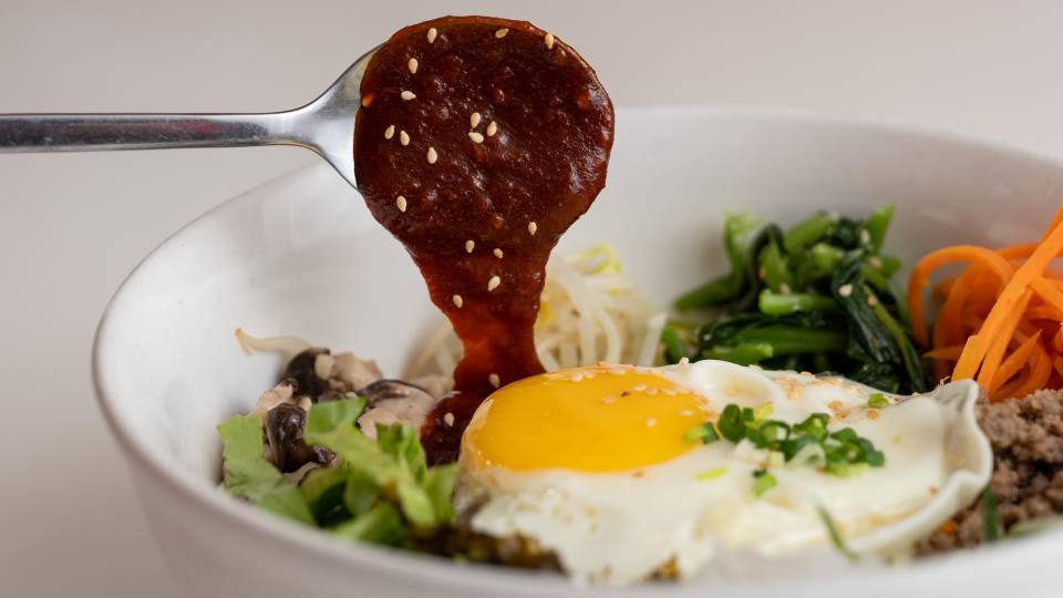 A bowl of bibimbap with mixed vegetables, a sunny-side up egg, and a spoon lifting sauce