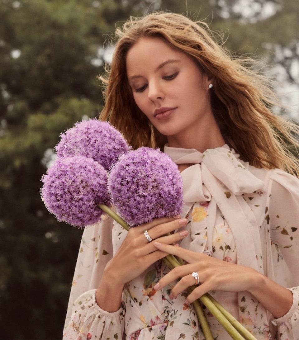 A woman with two engagement rings on her fingers poses holding a bouquet of flowers.