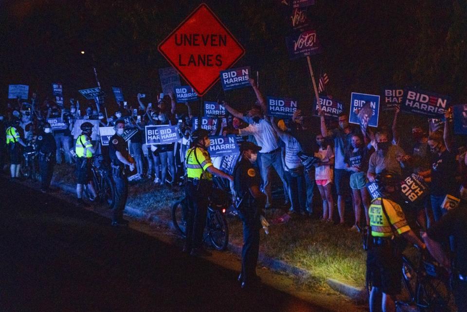 People with Biden-Harris campaign signs line a roadside as a handful of police officers stand in front of them