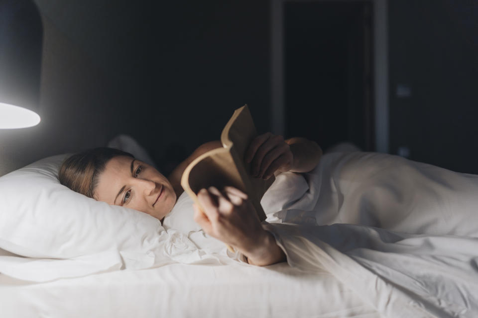 Person reading a book in bed with a lamp on, wrapped in white sheets