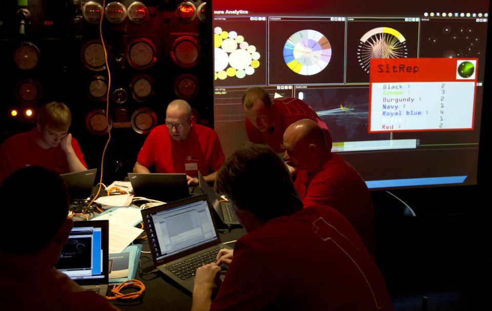 The 'Red team' work on their laptop computers next to a screen showing all teams progress during a mock cyberattack scenario with teams of amateur computer experts taking part and trying to fight this simulated attack in London, Friday, March, 14, 2014. The exercise comes complete with sirens and mock newscasts. It's meant to recruit the next generation of tech talent, and is also meant to help highlight the threat many here see as inevitable: A major cyberattack on the nation's critical infrastructure. (AP Photo/Alastair Grant)