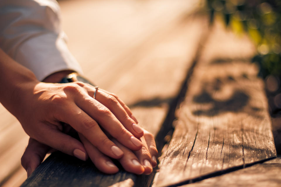 Two hands with intertwined fingers rest on a wooden surface. The visible hand sports an engagement ring. No faces are shown in the image