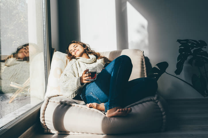 Woman relaxing in a sunlit room on a floor cushion, holding a cup, with a reflective surface nearby