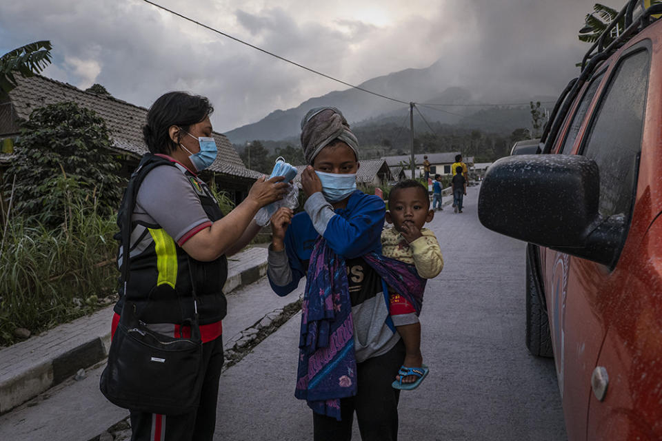 An official wearing a uniform of gray, navy, and red, helps a woman wearing a headdress with a child attached accross her back put on a blue mask