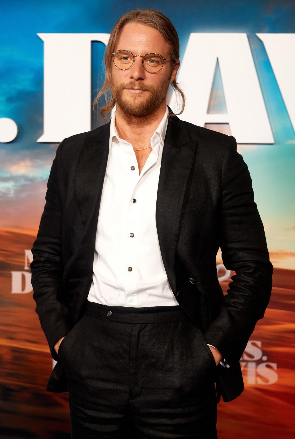 long hair tied back in a ponytail with a short beard and mustache and wearing glasses and a suit