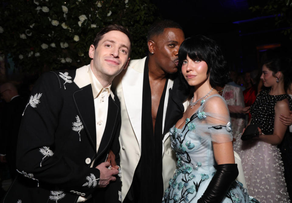 Three people posing close together; two in suits with embroidery, one in a blue floral dress