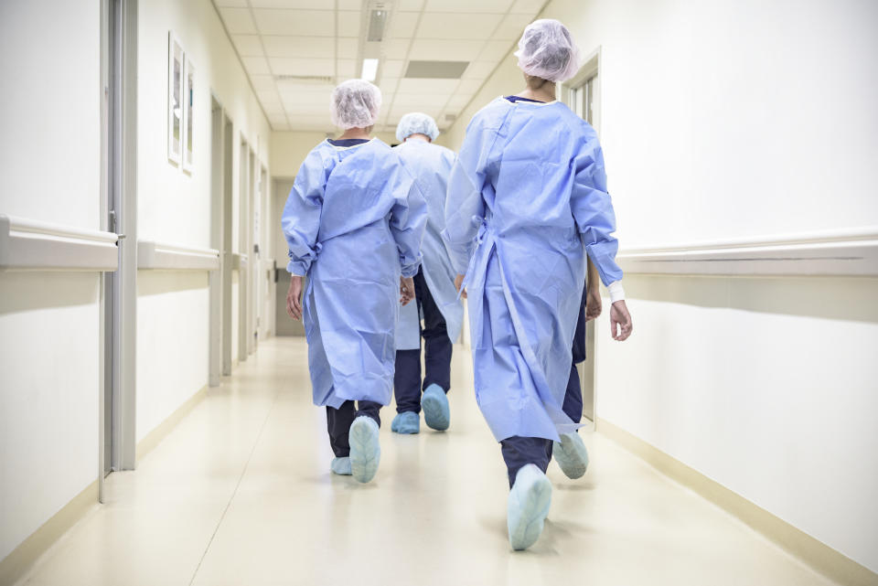 Rear view of male and female surgical team wearing gowns, caps, shoe covers, and walking down hospital corridor to operating room.