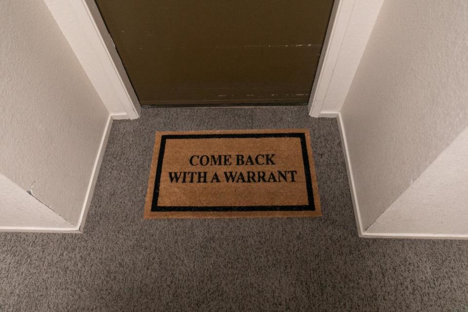 A doormat in front of a door has the words "Come back with a warrant" on it.