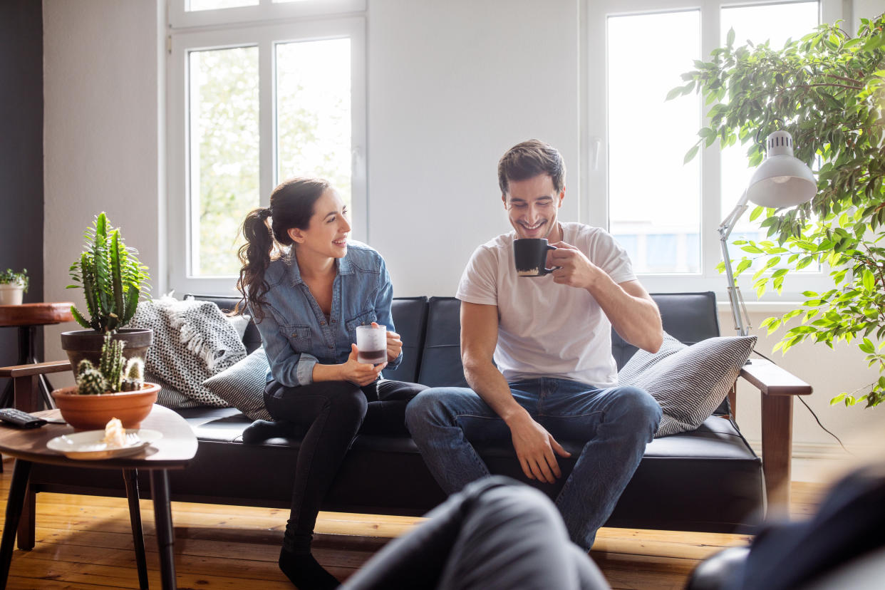 Cheerful young man and woman sitting on sofa in living room having coffee and talking