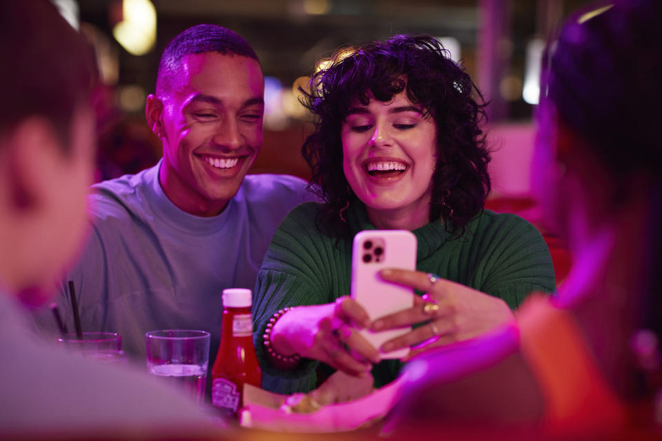 Cheerful Gen Z woman taking selfie with man on smart phone by friends at bowling alley