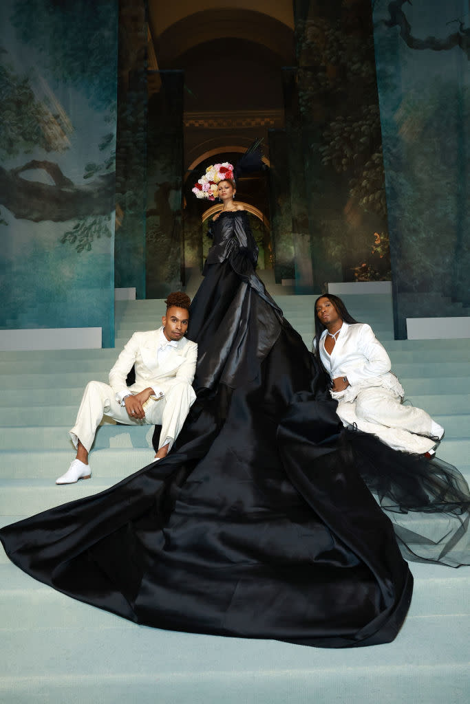 Two individuals sit beside a third person standing in an elaborate black gown with a floral headpiece, posed at an event