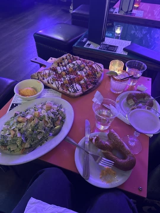 Assorted dishes on a table including a salad, a grilled platter, pretzel with dip, and beverages, viewed from a diner's perspective