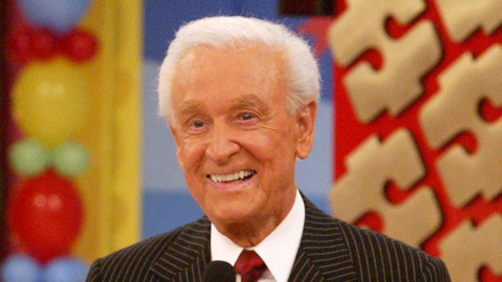 bob barker smiles while holding a microphone, he stands in front of a tv set with multicolored balloons and wears a black pinstriped suit jacket with a white collared shirt, red tie, and white pocket square