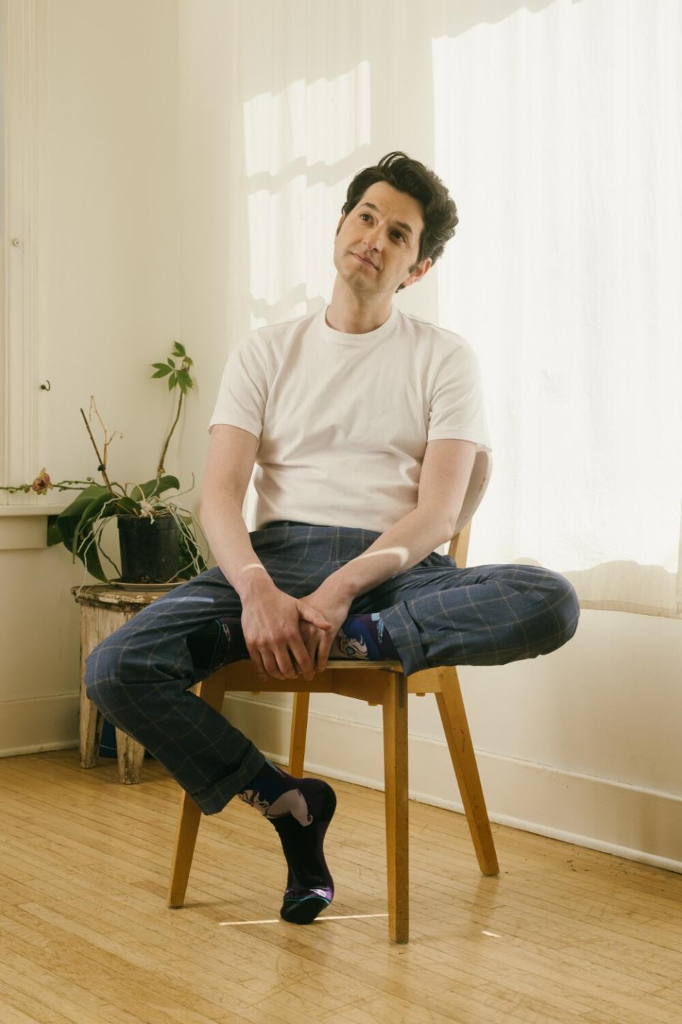 Ben Schwartz sits in a chair in front of white curtains.