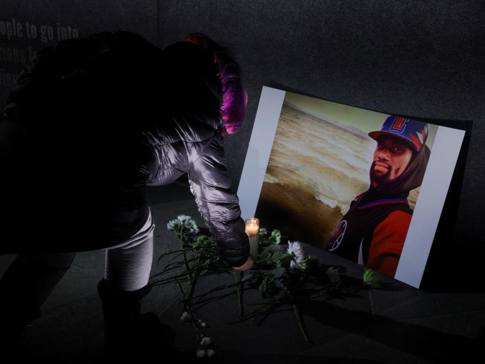 A woman in a Black jacket leaves white flowers in front of a photo of Tyre Nichols in front of a sunset