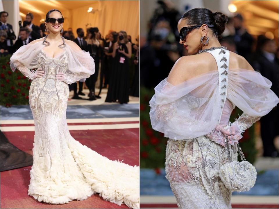 Side by side of the front and back of Rosalia's dress. It's pure white and off-white with a lot of sheer materials, texture, and embelishments. The sweetheart neckline is surrounded by tulle off-the-shoulder sleeves held up my metal hooks around her neck. The bodice is covered intricate detailing and is fitted until it flares out at the skirt with a ruffle and feather-like train. From the back, it shows a racerback with the tulle connected and more intricate beading around the bodice. She's wearing black sunglasses and holding a matching round fabric bag.