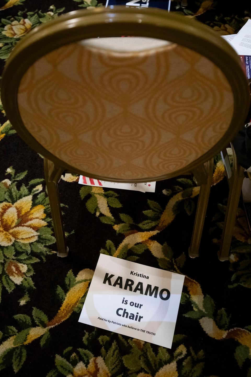 A sign on the floor under a chair says "Kristina Karamo is our chair, paid for by patriots who believe in the truth!"