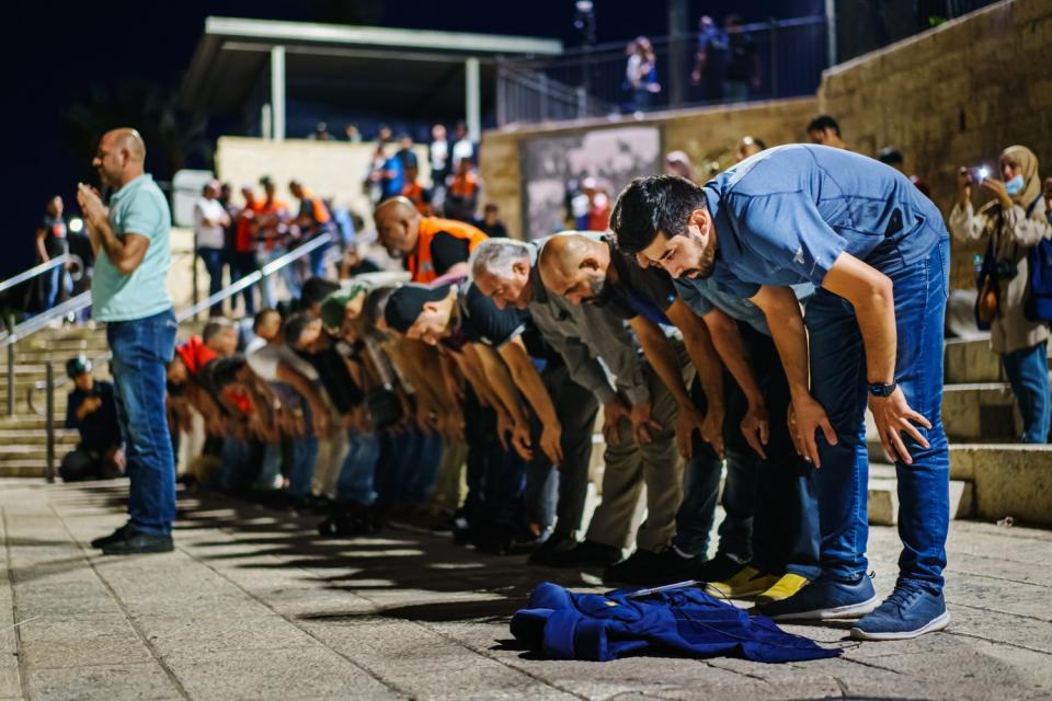 A man with hands folded in prayer stands in front of a line of men bending forward at the waist.