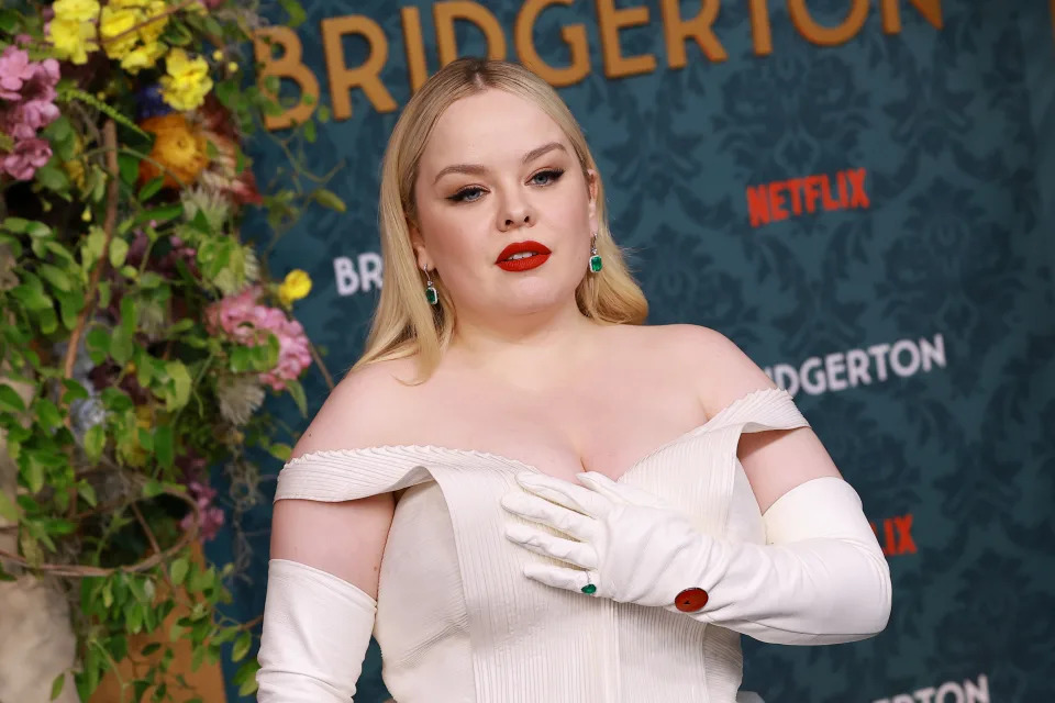 Nicola Coughlan at a Bridgerton event, wearing an off-shoulder dress with gloves and bold makeup and the pin on her glove