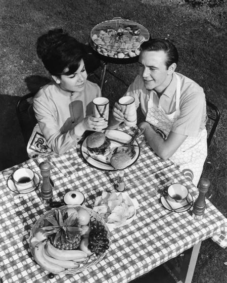 A woman and a man sitting over a meal at a table with a checkered tablecloth, a charcoal grill behind them