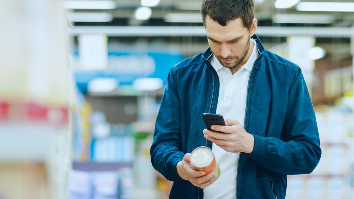 At the Supermarket: Handsome Man Uses Smartphone to Check Nutritional Value of the Canned Goods and Buy it.