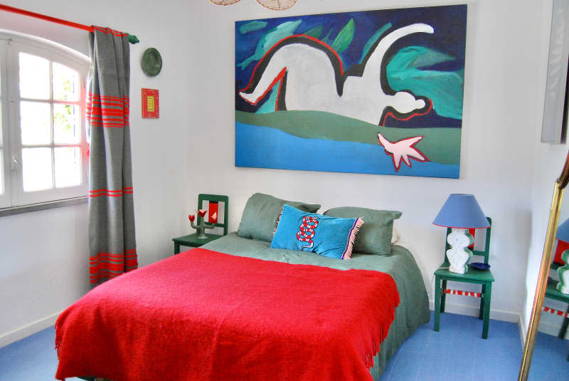Artwork hung above bed with green and red bedding in white bedroom with blue floor.