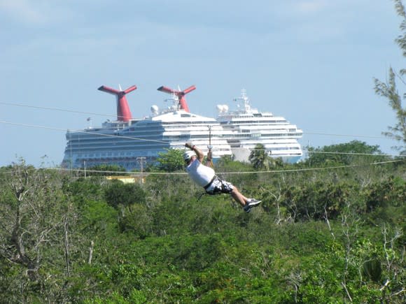 Person ziplining over a forest canopy with cruise ships in the background.